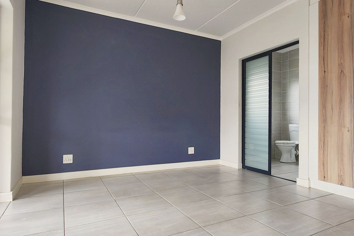 A bright bedroom featuring tiled flooring, a dark blue accent wall, built-in wooden cabinetry, and an en-suite bathroom with a sliding glass door.