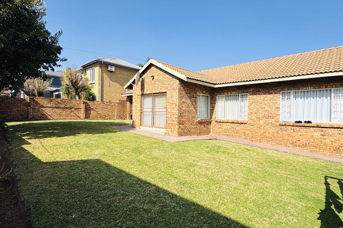 A well-maintained rear exterior view of a face brick house featuring a spacious lawn, paved walkway, and secure perimeter walling.