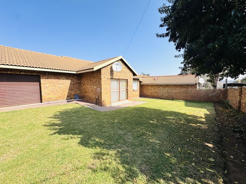 A rear view of the property featuring a face brick facade, tiled roof, roller garage door, and a paved patio area leading onto a lawn.