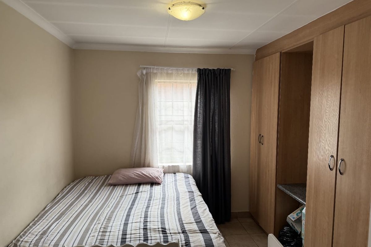 A functional bedroom featuring built-in wooden cupboards, tiled flooring, and a window with curtains, providing a practical living space.