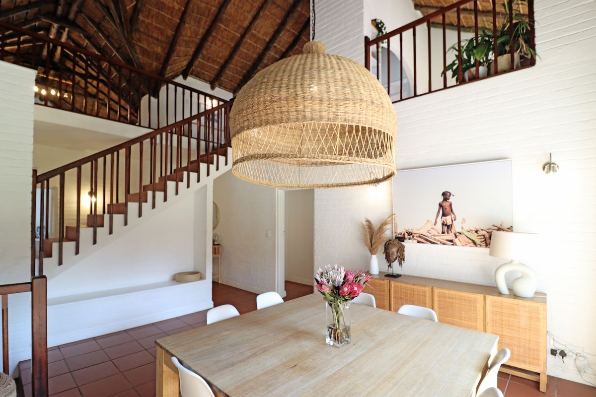 Spacious living area featuring a double-volume thatch ceiling, exposed timber rafters, a mezzanine level, and a classic timber staircase with terracotta flooring.