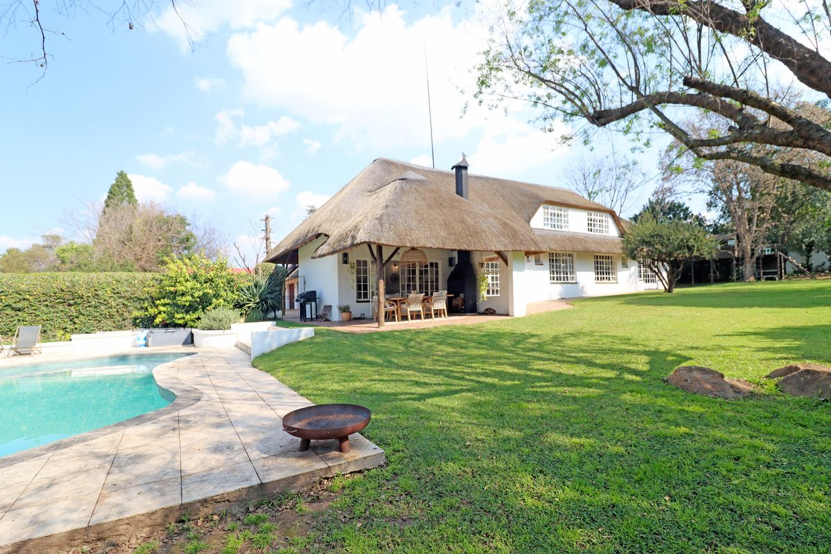 Rear view of a charming thatched-roof home featuring a covered patio, outdoor fireplace, swimming pool, and expansive landscaped garden.