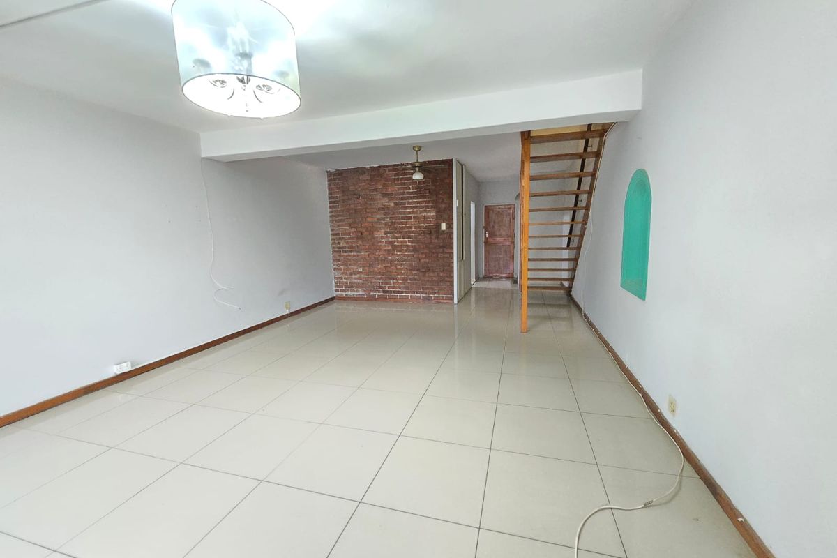 An empty, open-plan living area featuring tiled floors, an exposed brick feature wall, and a wooden staircase leading to the upper level.