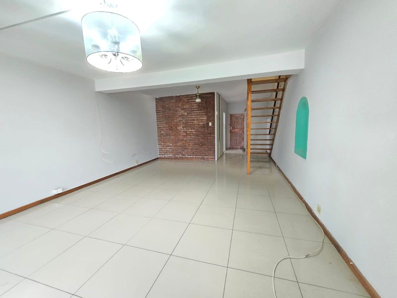 An empty, open-plan living area featuring tiled floors, an exposed brick feature wall, and a wooden staircase leading to the upper level.