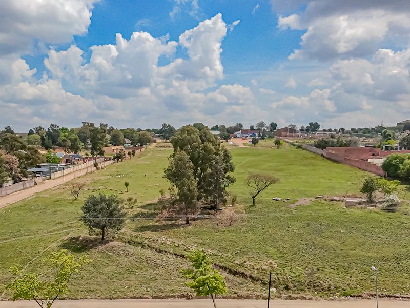 An aerial view of a large, vacant residential plot featuring open grass land, scattered trees, and surrounding boundary walls in a suburban setting.
