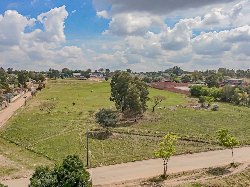 An aerial view of a large, vacant residential plot featuring open grass, scattered trees, and surrounding neighborhood infrastructure.