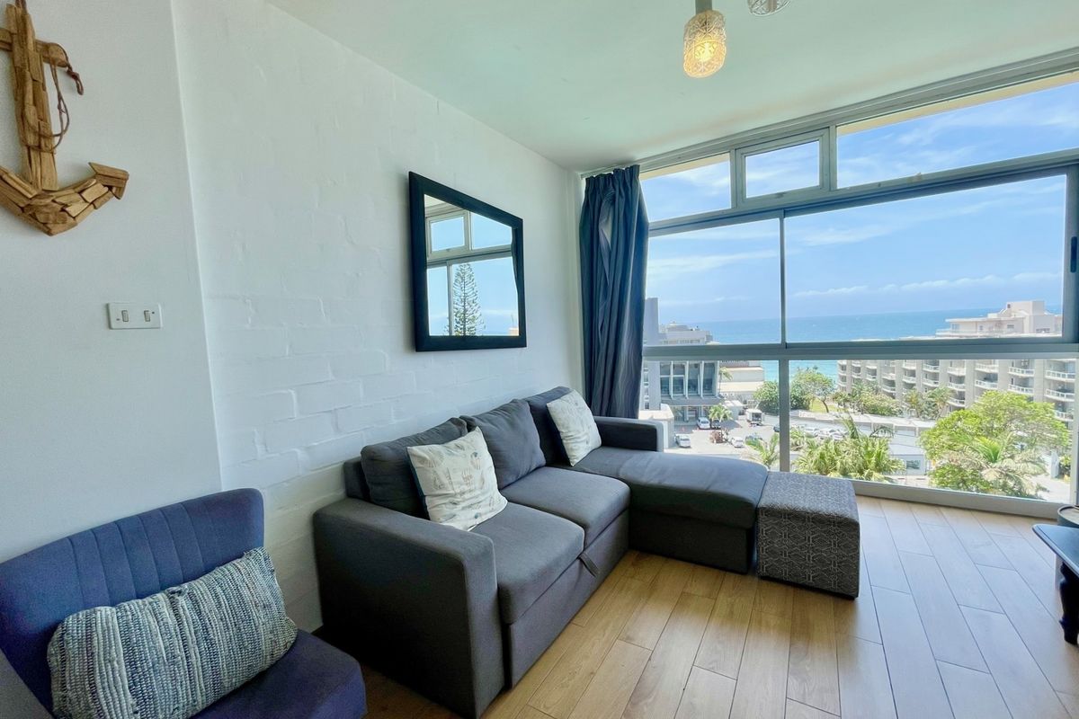 A bright living room featuring large floor-to-ceiling windows with expansive ocean views, wood-look flooring, and a painted brick feature wall.