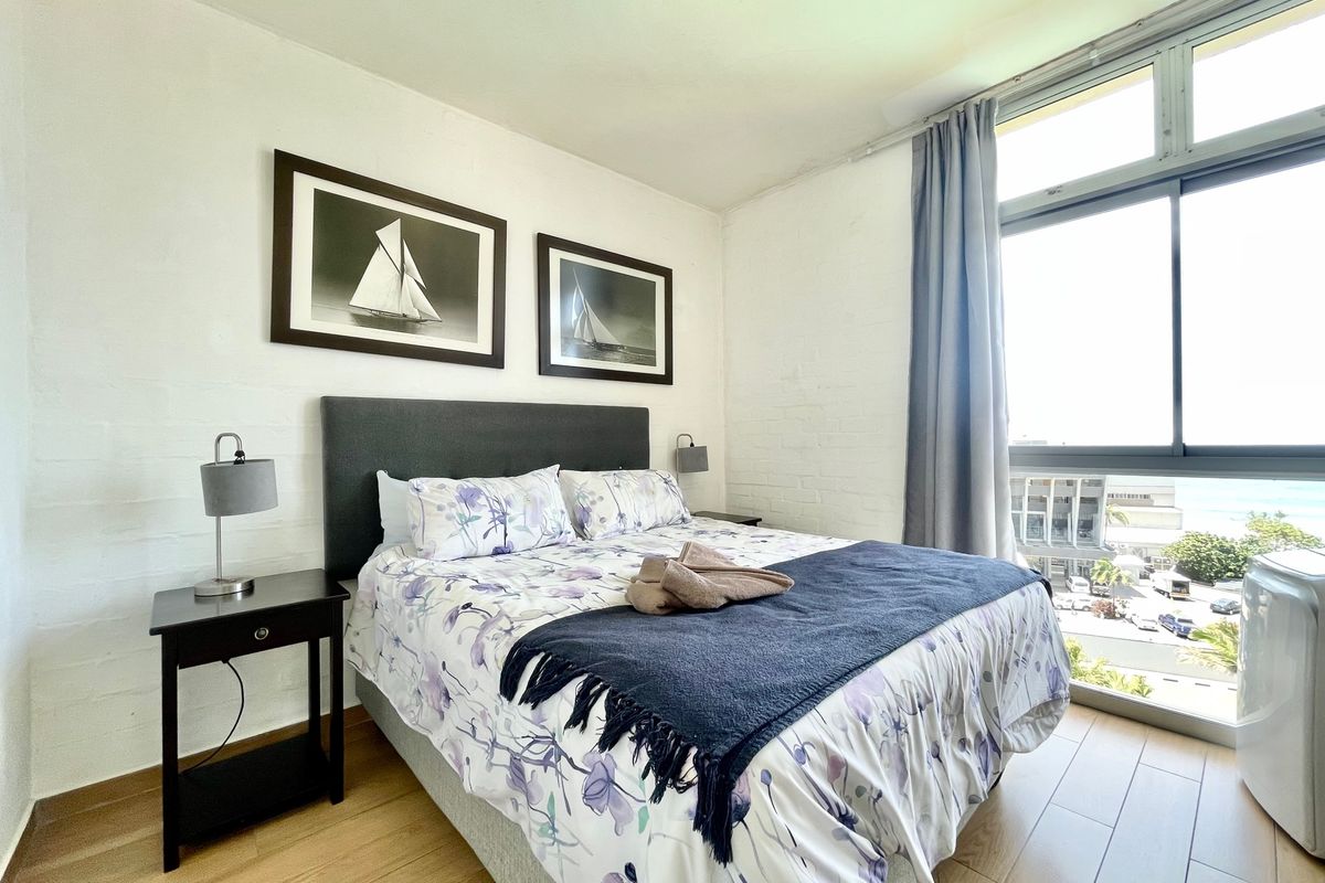A bright bedroom featuring light wood-look flooring, white painted brick walls, and a large window providing natural light and an exterior view.