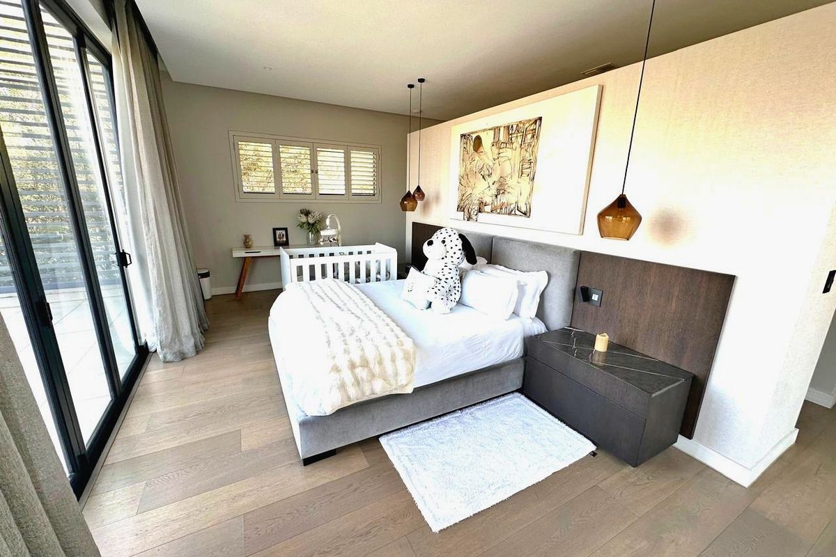 A modern, well-appointed bedroom featuring timber flooring, integrated bedside cabinetry, pendant lighting, and large sliding glass doors opening to an outdoor area.