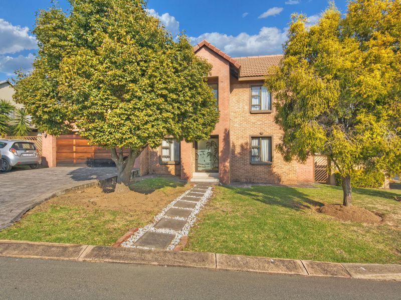 A well-maintained double-story brick house featuring a paved driveway, double garage with wooden doors, and a landscaped front garden with a paved walkway.