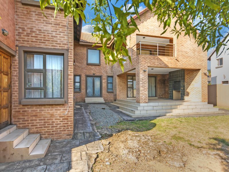 Rear view of a double-storey face brick house featuring a covered patio with a built-in braai, an upper balcony, and paved outdoor areas.