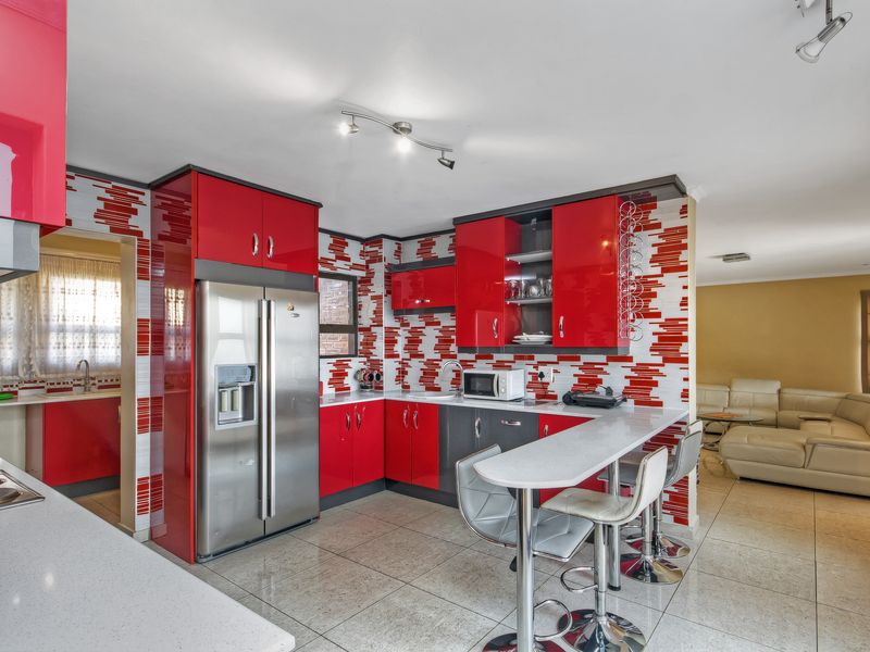 Modern kitchen featuring vibrant red high-gloss cabinetry, stone countertops, a breakfast bar, and distinctive mosaic wall tiling, seamlessly connected to the living area.