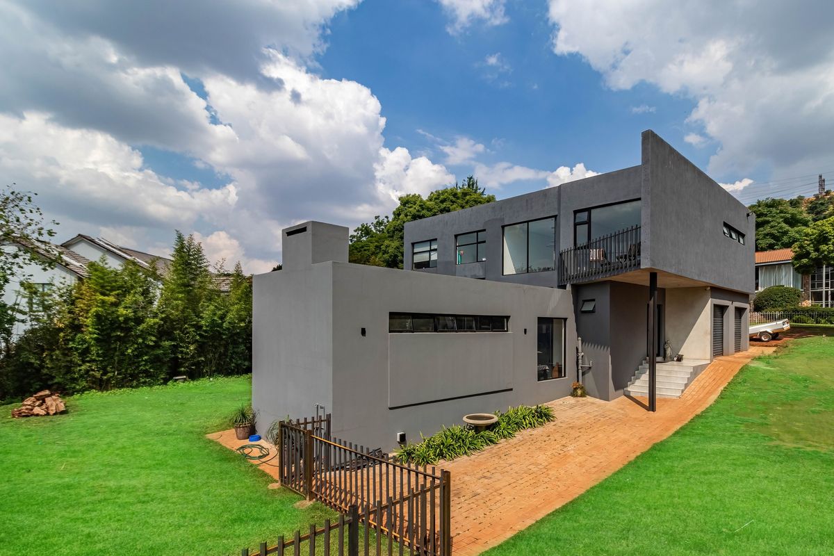 A modern two-story house with a grey exterior, featuring a balcony, large windows, and a brick-paved walkway set within a spacious green garden.