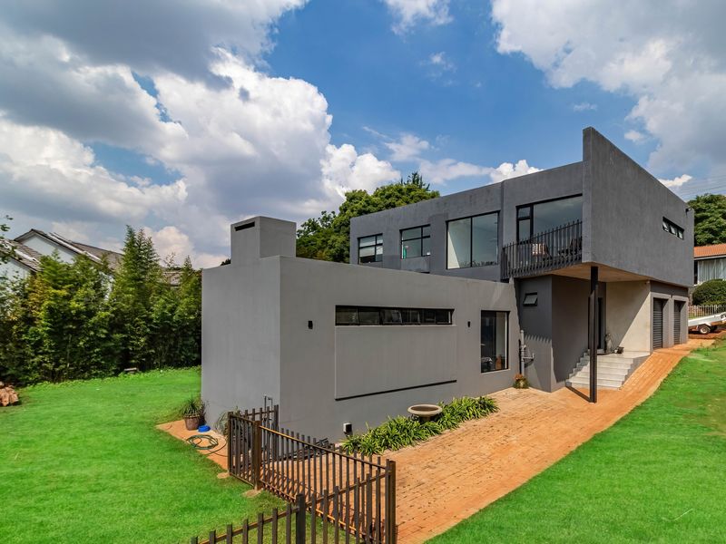 A modern two-story house with a grey exterior, featuring a balcony, large windows, and a brick-paved walkway set within a spacious green garden.