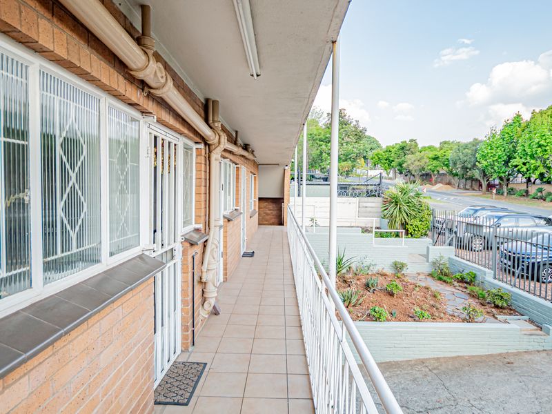 An exterior walkway or balcony featuring brick walls, tiled flooring, and metal security railings, overlooking a small garden area and street parking.