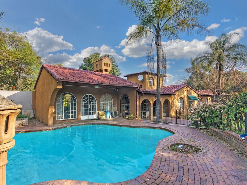 A spacious outdoor pool area featuring a curved swimming pool, brick paving, and a Mediterranean-style home with arched windows and terracotta roof tiles.