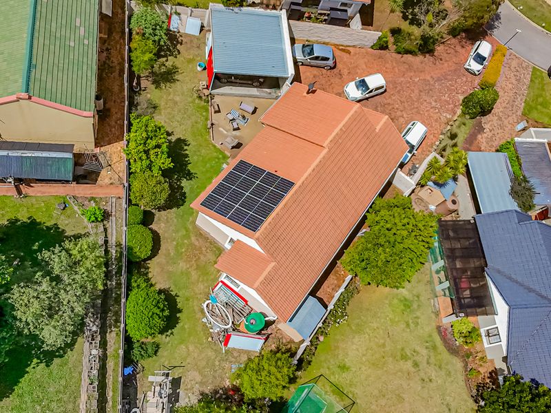 Aerial view of the property featuring a tiled roof with solar panels, a detached garage, paved driveway, and surrounding garden space.