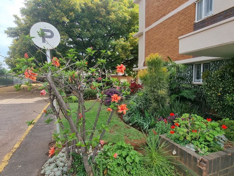 Front exterior view of the flat building featuring a brick facade, concrete balcony, and a well-maintained landscaped garden bed with a brick retaining wall.