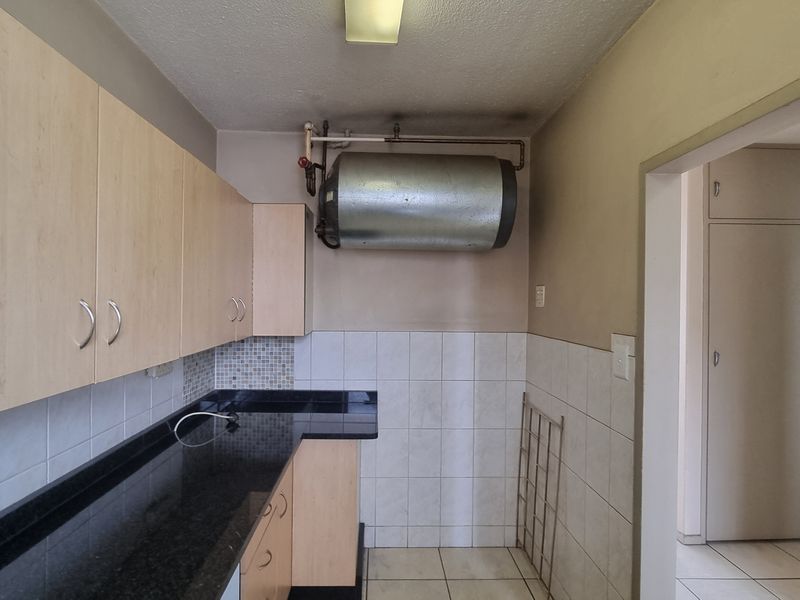 A compact kitchen featuring laminate cabinetry, granite countertops, and tiled splashbacks, with an exposed wall-mounted water heater.