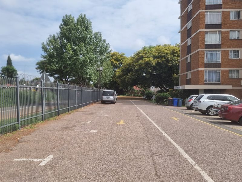 Exterior view of the apartment complex showing the parking area, paved driveway, and perimeter security fencing.