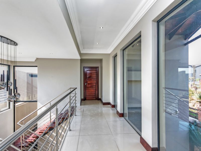 A bright, modern upper-level hallway featuring polished tile flooring, a sleek stainless steel balustrade, and large windows overlooking the exterior.
