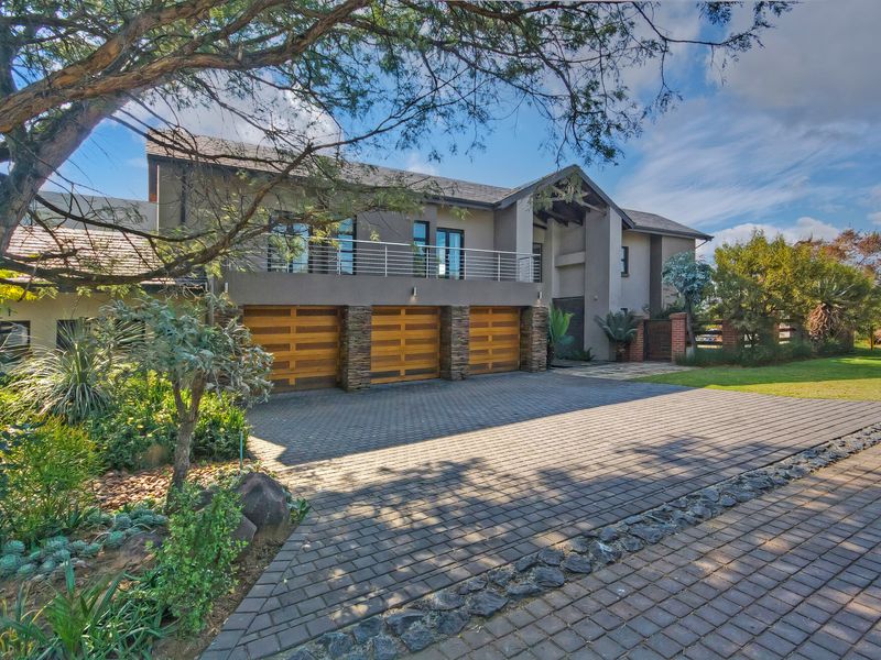 A modern double-story residence featuring a triple garage, paved driveway, stone-clad accents, and an upper-level balcony overlooking a well-maintained landscaped garden.