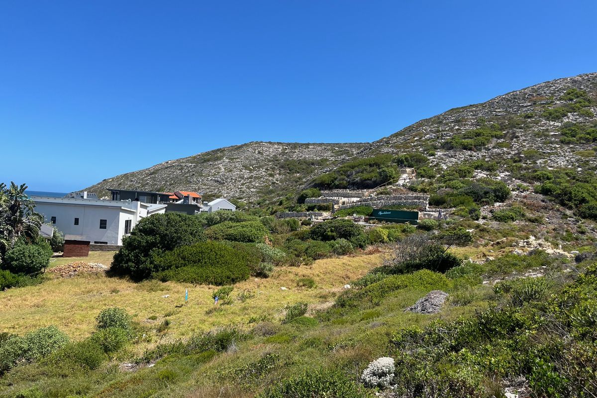 A view of the rear residential plot featuring terraced stone retaining walls and natural coastal vegetation set against a hillside backdrop.