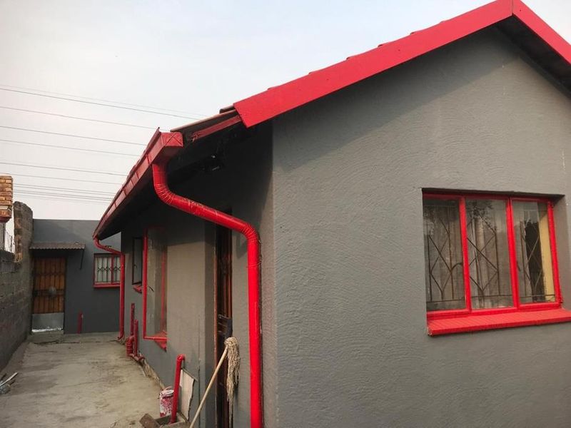 Rear exterior view of the house featuring grey rendered walls, contrasting red roof trim, window frames, and downpipes with security bars on windows.