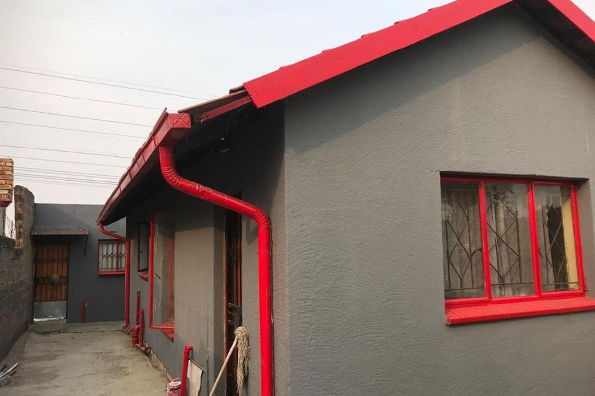 Rear exterior view of the house featuring grey rendered walls, contrasting red roof trim, window frames, and downpipes with security bars on windows.
