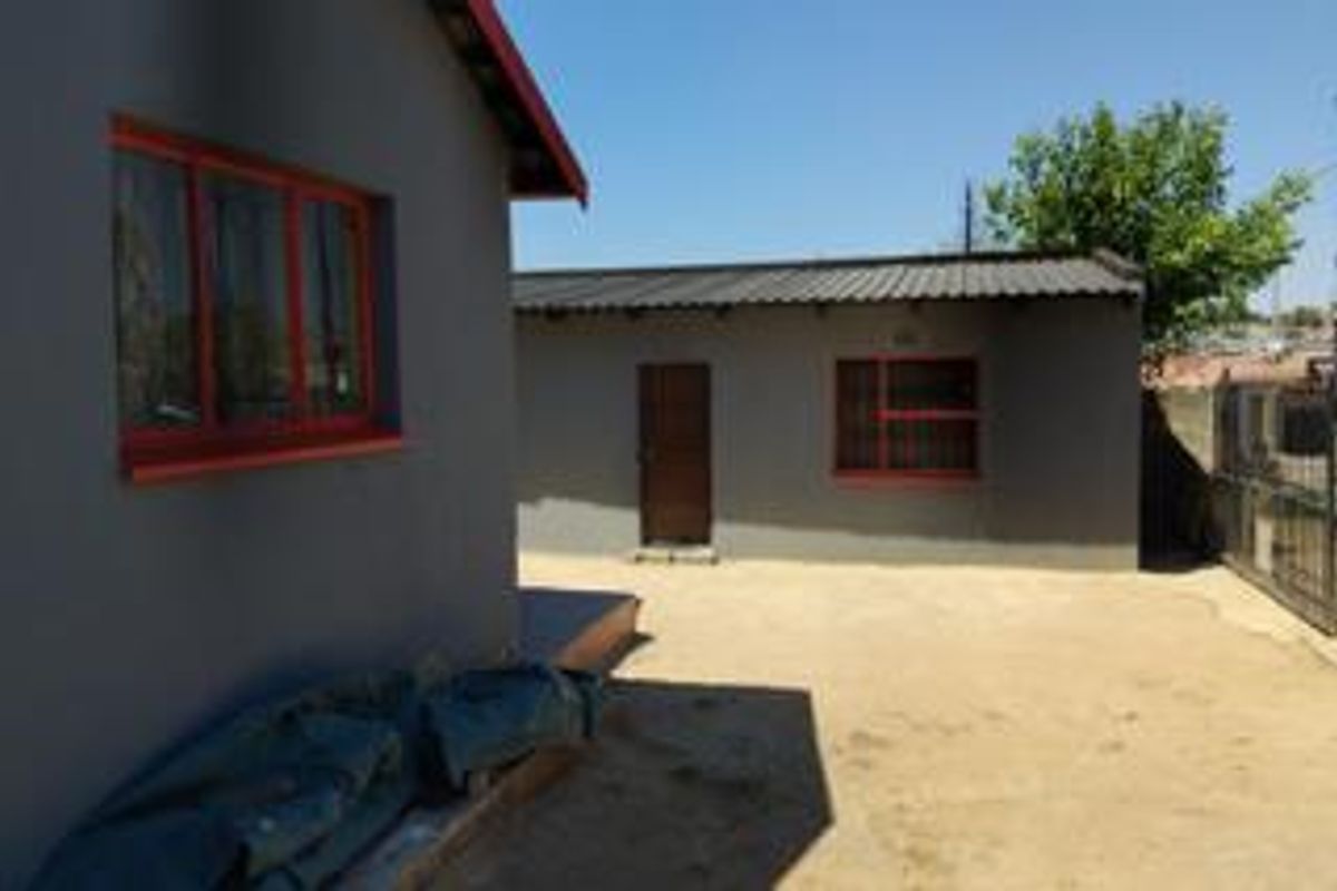 Rear exterior view of the property featuring grey painted walls, red steel window frames, and a metal roof with a concrete yard area.