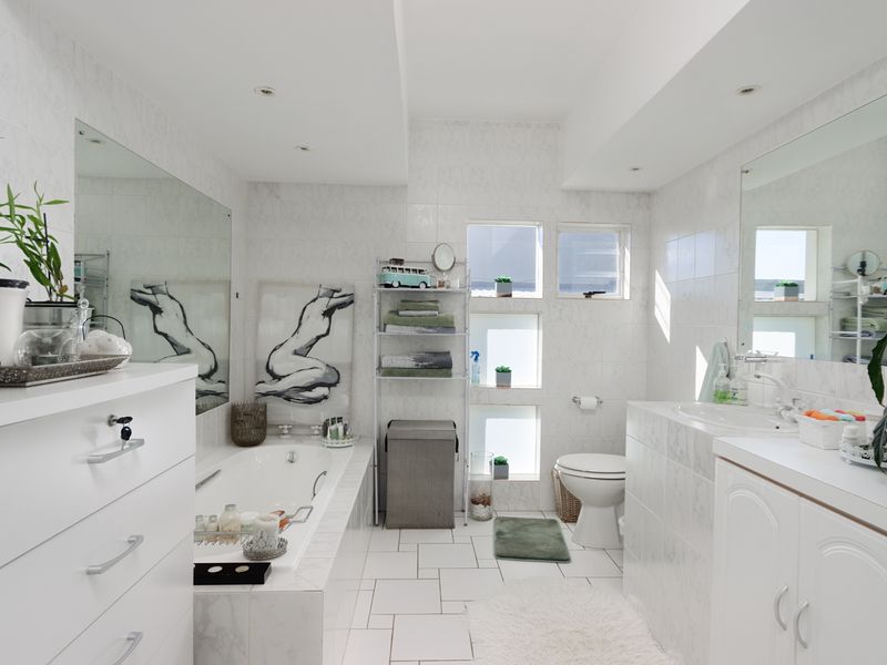 A bright, fully tiled bathroom featuring a built-in bathtub, vanity cabinet with integrated sink, and large wall-mounted mirrors.