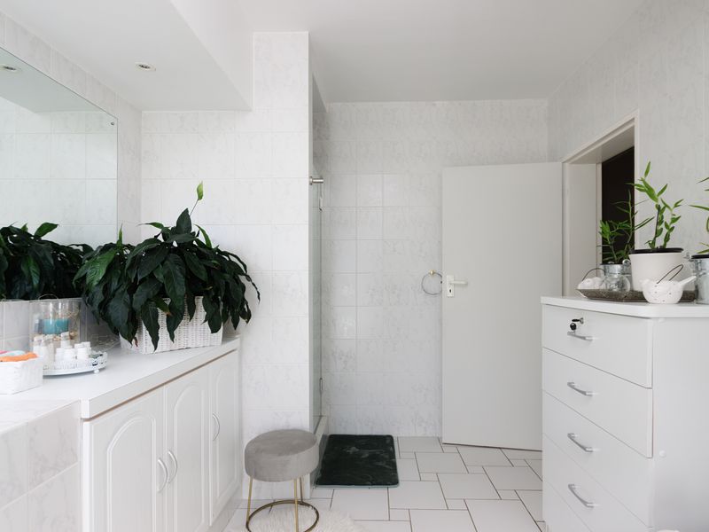 A bright bathroom featuring white tiled walls and flooring, a built-in vanity cabinet with a large mirror, and a separate shower enclosure.