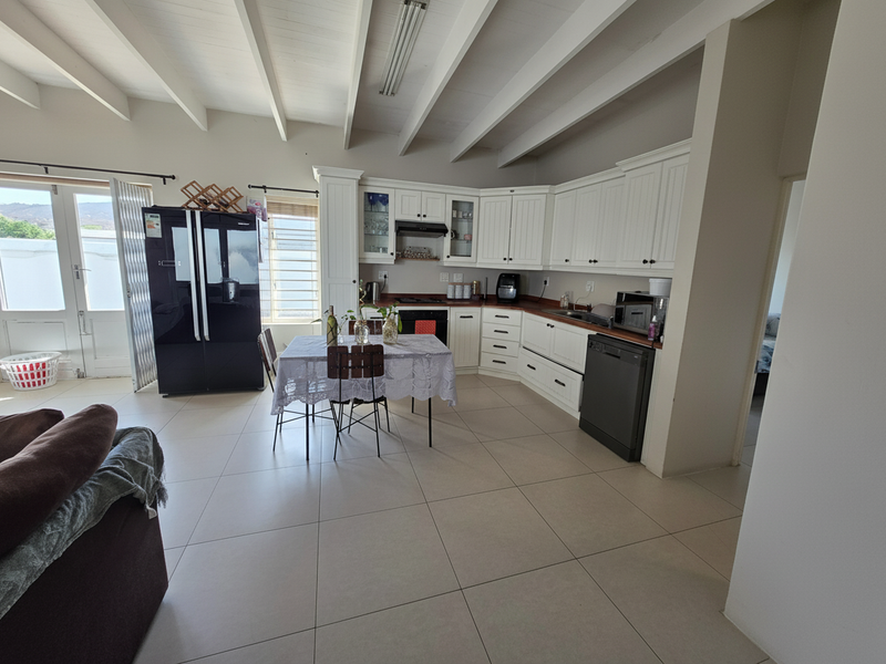 Spacious open-plan kitchen and dining area featuring white cabinetry, dark countertops, exposed beam ceilings, and large tiled flooring with direct access to the patio.
