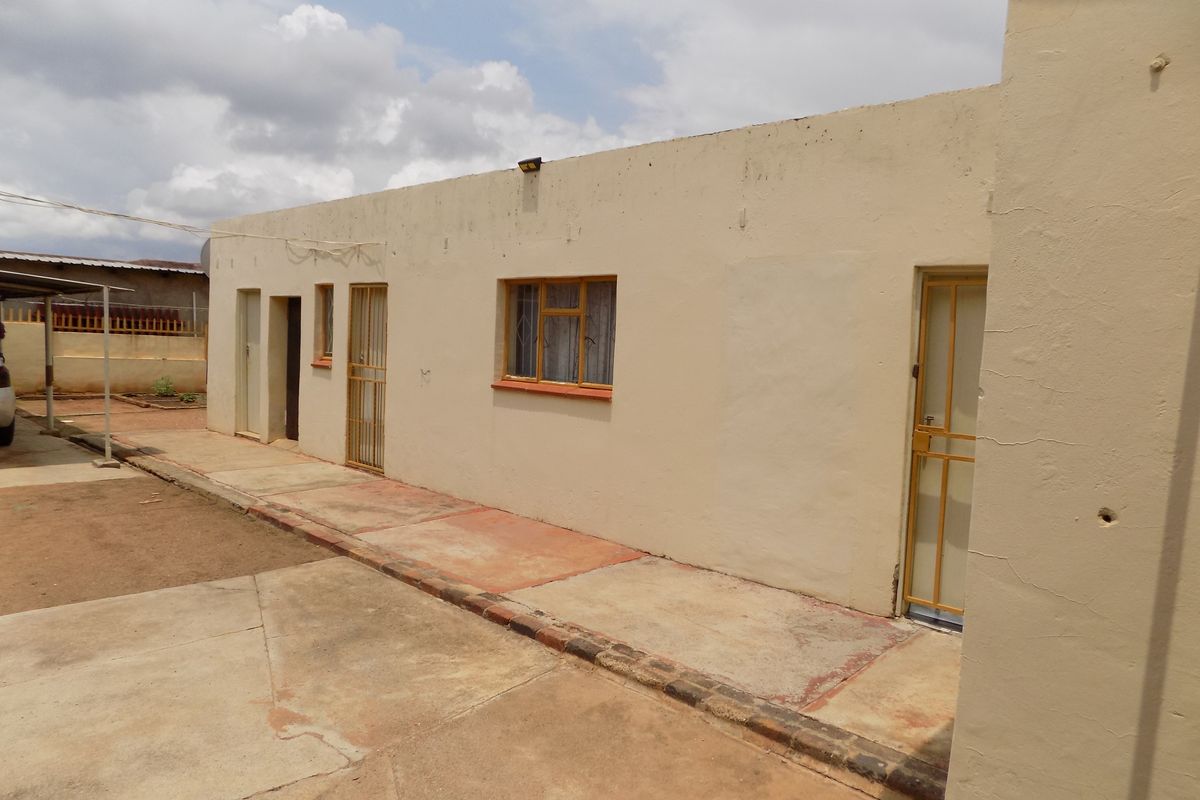 Rear exterior view of the property featuring concrete paving, security gates, and metal window frames, showing signs of wear and weathering.