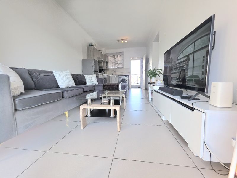 An open-plan living area featuring tiled flooring and a compact kitchen with built-in cabinetry and laundry facilities, leading to an outdoor balcony.