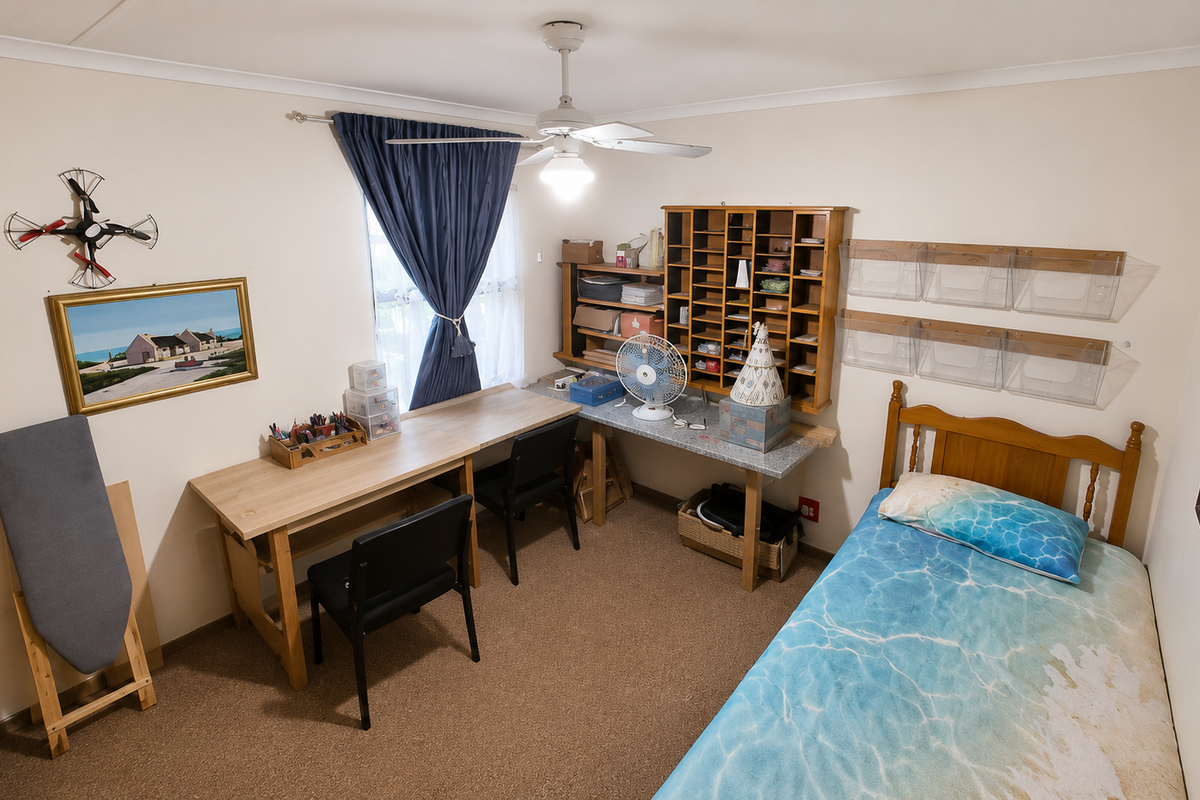 A functional bedroom featuring a ceiling fan, built-in wall shelving, and carpeted flooring, currently utilized as a combined sleeping and workspace area.