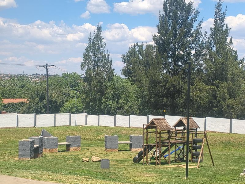 Spacious communal garden area featuring a perimeter wall, built-in brick braai facilities, and a wooden playground set for outdoor recreation.