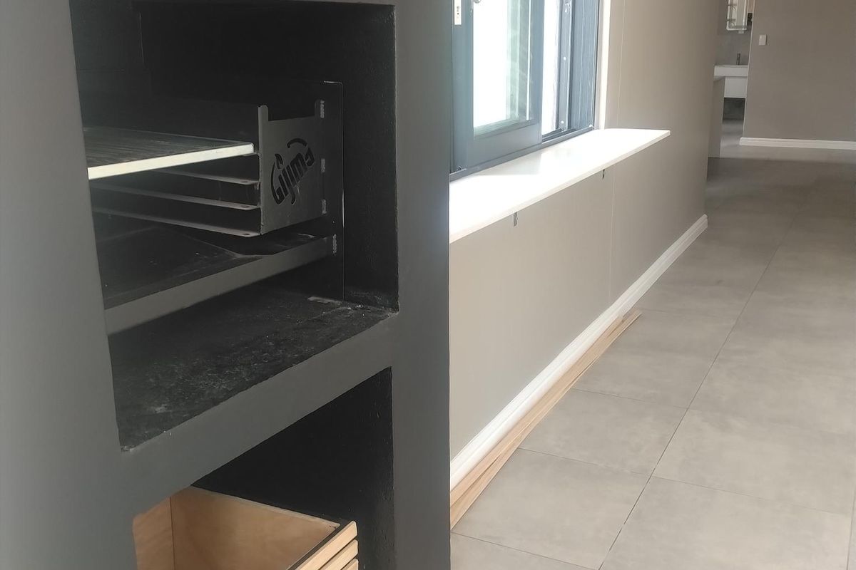 An open-plan living area featuring a built-in braai, modern grey floor tiles, and a sliding window with a white sill.