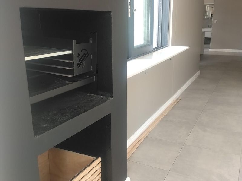 An open-plan living area featuring a built-in braai, modern grey floor tiles, and a sliding window with a white sill.