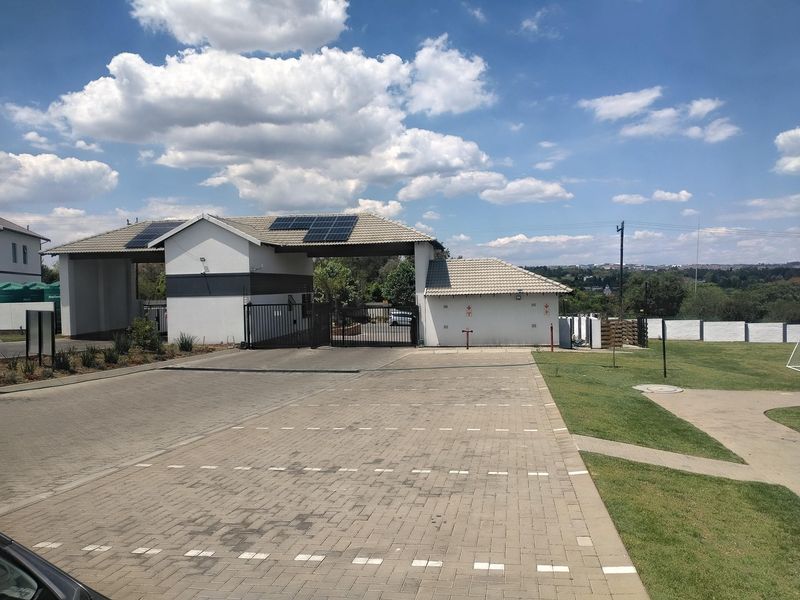 A well-maintained residential complex entrance featuring a security gate, guard house, paved driveway, and solar panel installation on the roof.