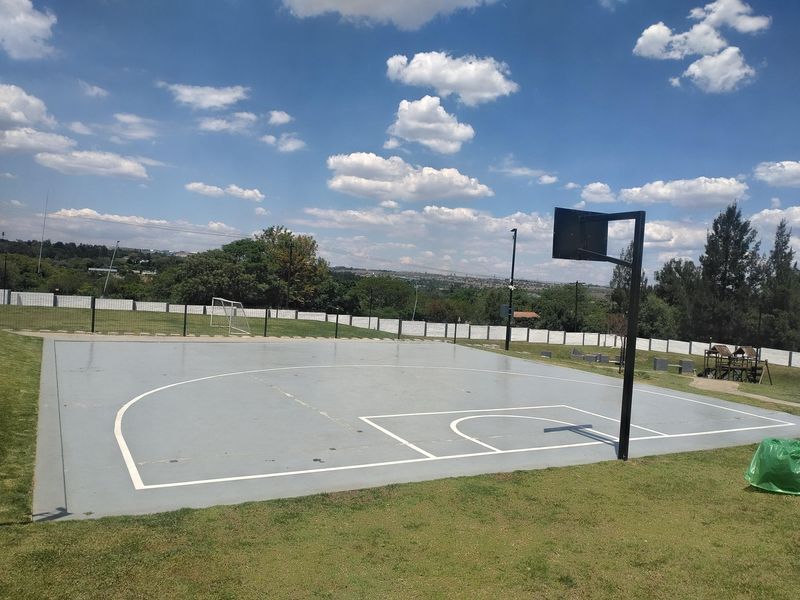 Outdoor sports facility featuring a paved basketball court with a hoop, surrounded by landscaped lawn and perimeter fencing.