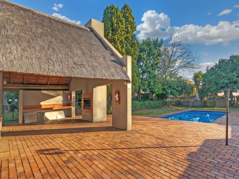 Outdoor entertainment area featuring a covered patio with a thatched roof, built-in braai, brick paving, and a swimming pool set in a garden.