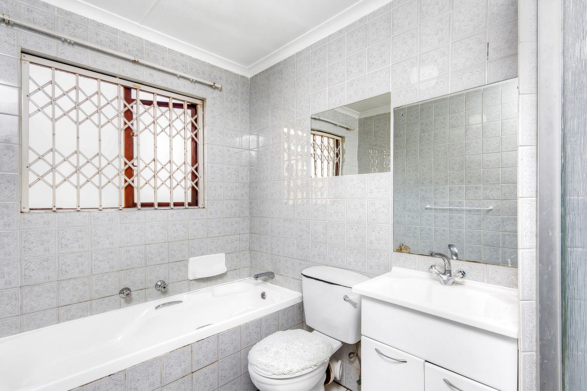 A functional bathroom featuring a built-in bathtub, vanity cabinet, and full-height ceramic wall tiling, equipped with a security gate on the window.
