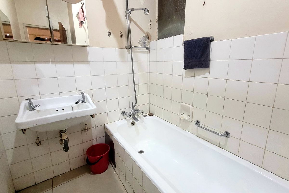A dated bathroom featuring a wall-mounted basin, bathtub with shower mixer, and tiled walls, currently requiring maintenance and cosmetic updates.