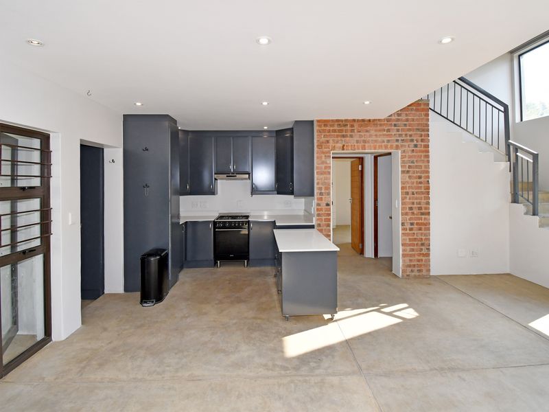 Modern open-plan kitchen featuring dark cabinetry, a central island, and exposed brick accents, seamlessly integrated with a staircase in a spacious, light-filled living area.