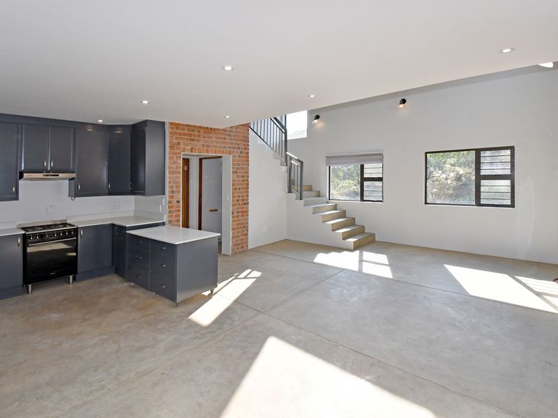 Spacious open-plan living area featuring a modern kitchen with dark cabinetry, concrete floors, exposed brick accents, and a staircase leading to the upper level.