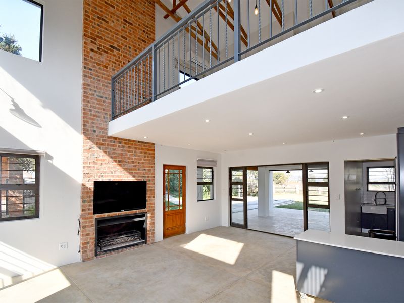 Spacious open-plan living area featuring a double-volume ceiling, exposed brick fireplace, mezzanine level, and seamless flow to the outdoor patio.