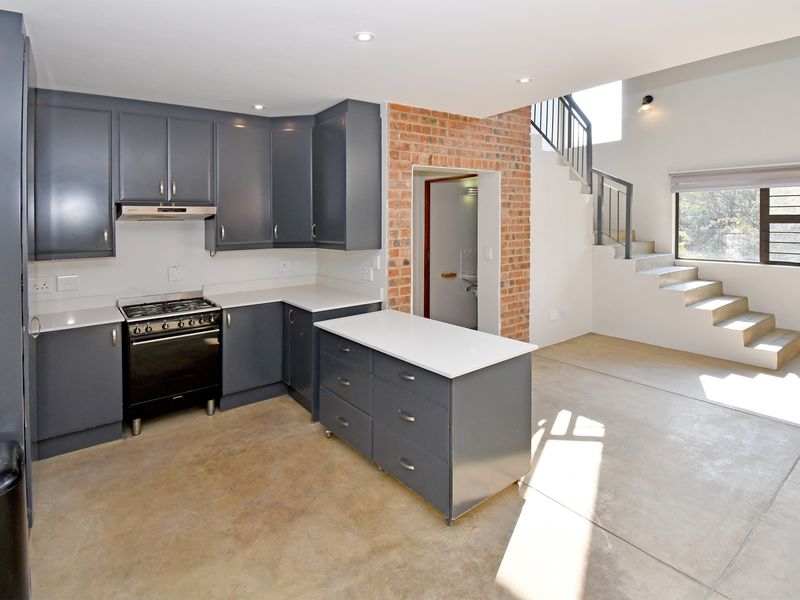 Modern open-plan kitchen featuring dark cabinetry, a central island, and exposed brickwork, seamlessly connected to a staircase area with polished concrete flooring.