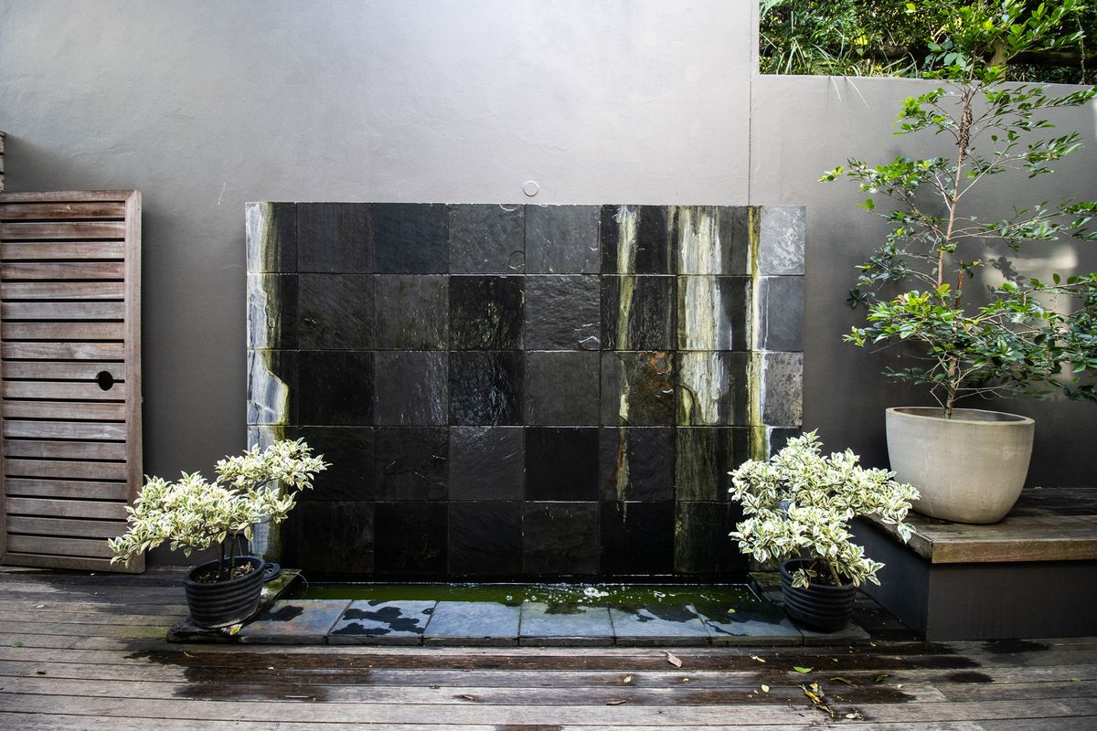 A rear outdoor area featuring a dark tiled water feature set against a grey wall with timber decking.