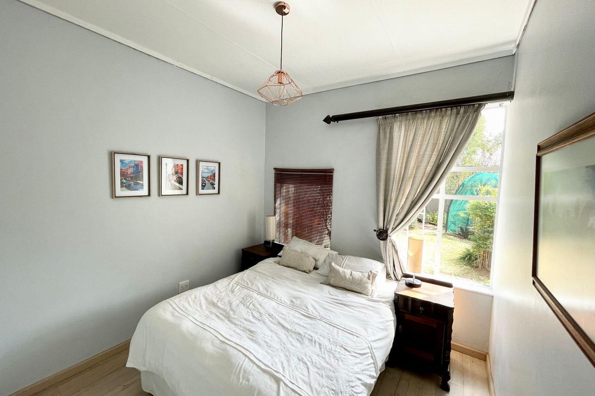 A well-lit bedroom featuring light-toned laminate flooring, a large window with curtain rod, and a modern geometric pendant light fixture.
